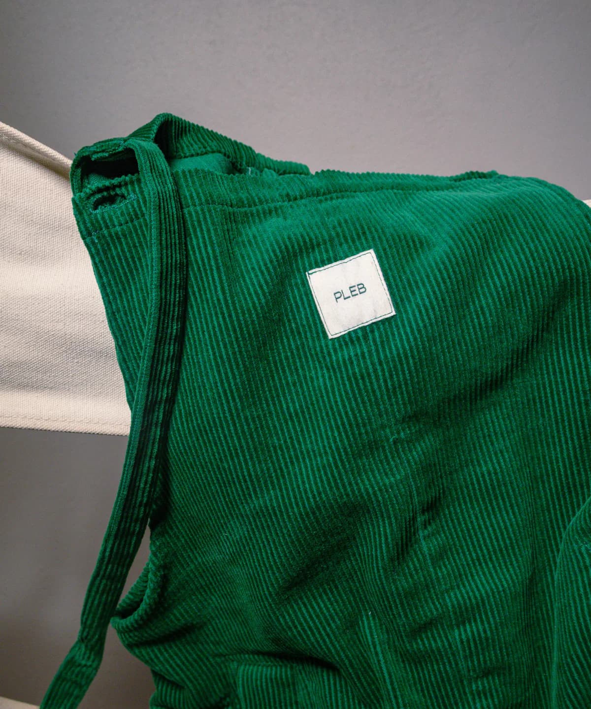 Forest green PLEBS cotton corduroy dungarees draped over a light chair, showing the mid-wale rib texture and woven PLEBS label.
