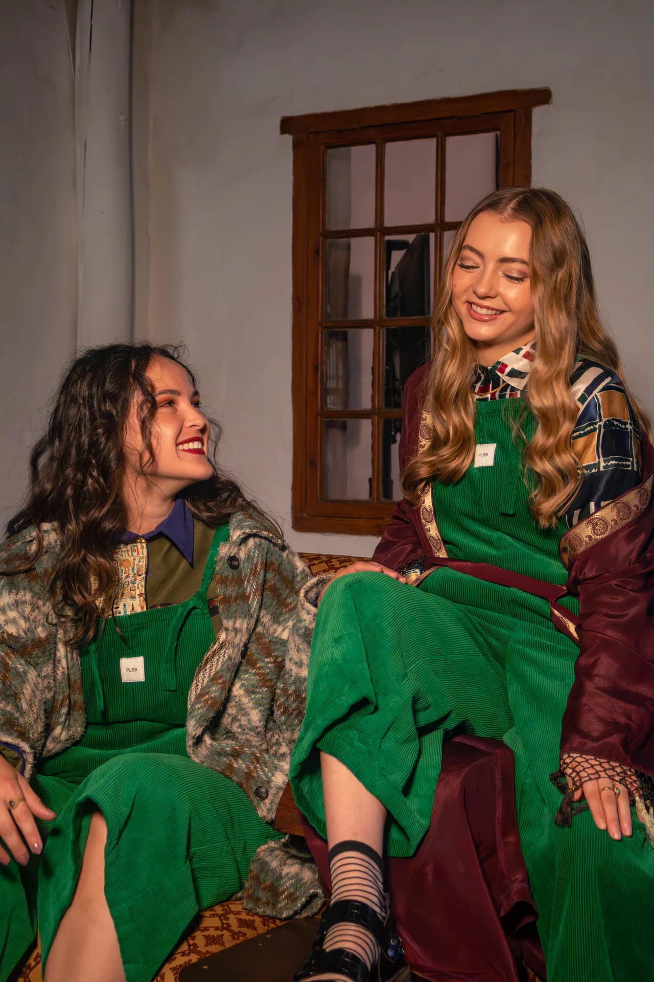 Two people sitting indoors in forest green PLEBS cotton corduroy dungarees, styled with layered jackets.