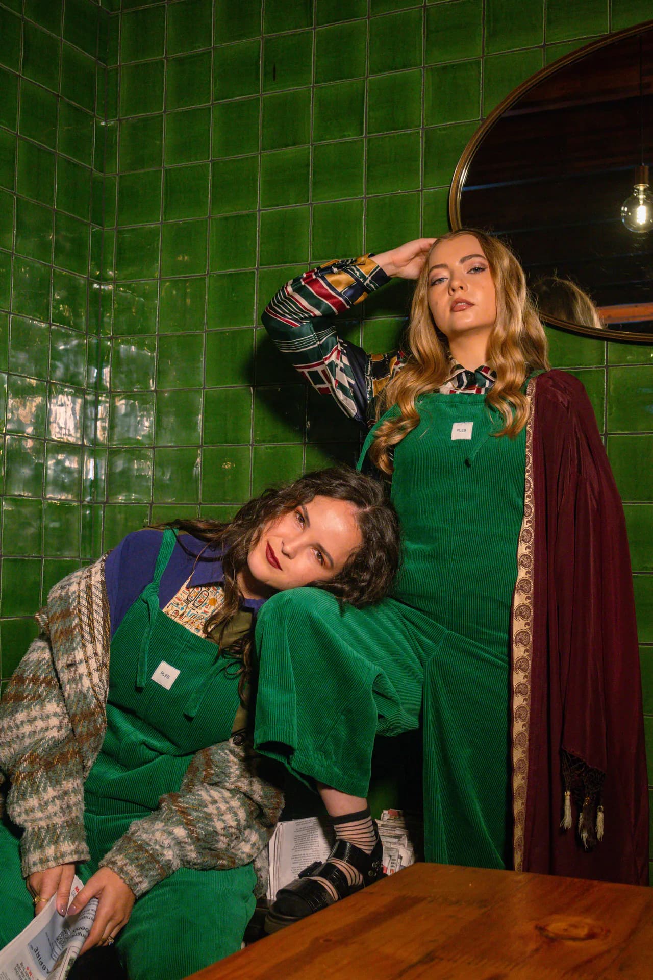 Two people posing against green tiles in forest green PLEBS cotton corduroy dungarees.
