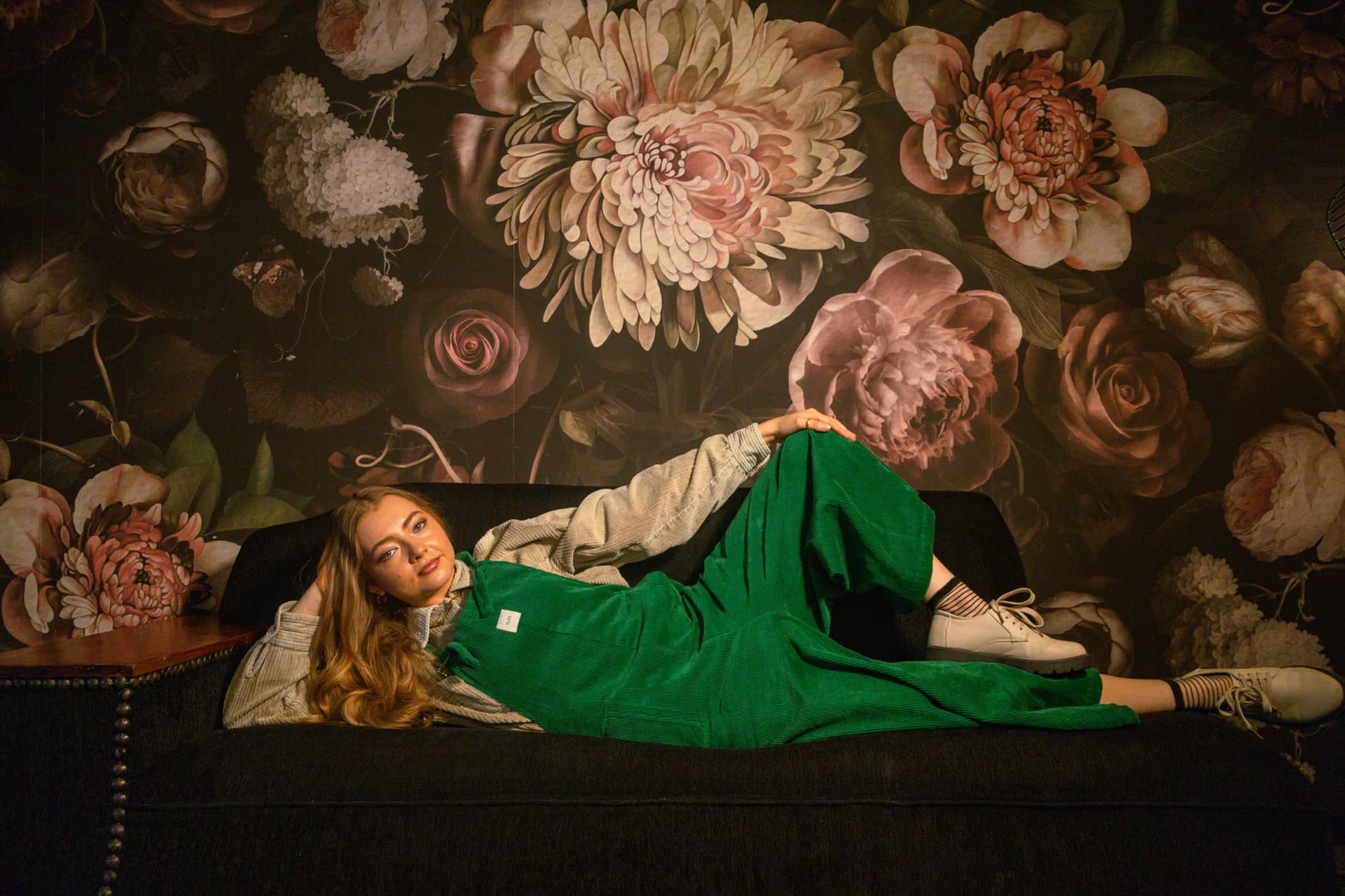 Person reclining on a sofa in forest green PLEBS cotton corduroy dungarees over a light shirt.