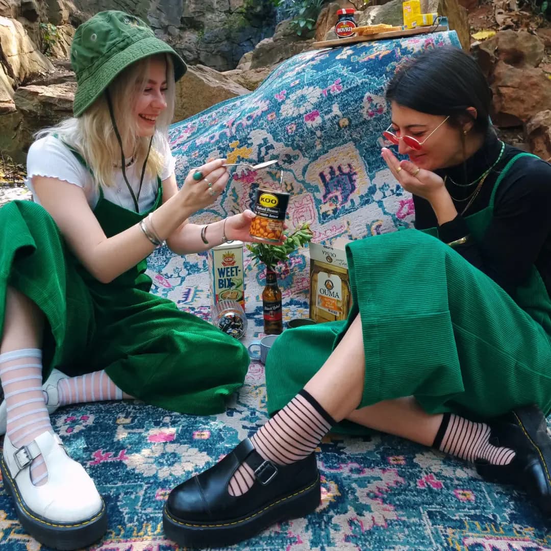 Two people wearing green PLEBS cotton corduroy dungarees seated together at an outdoor picnic.