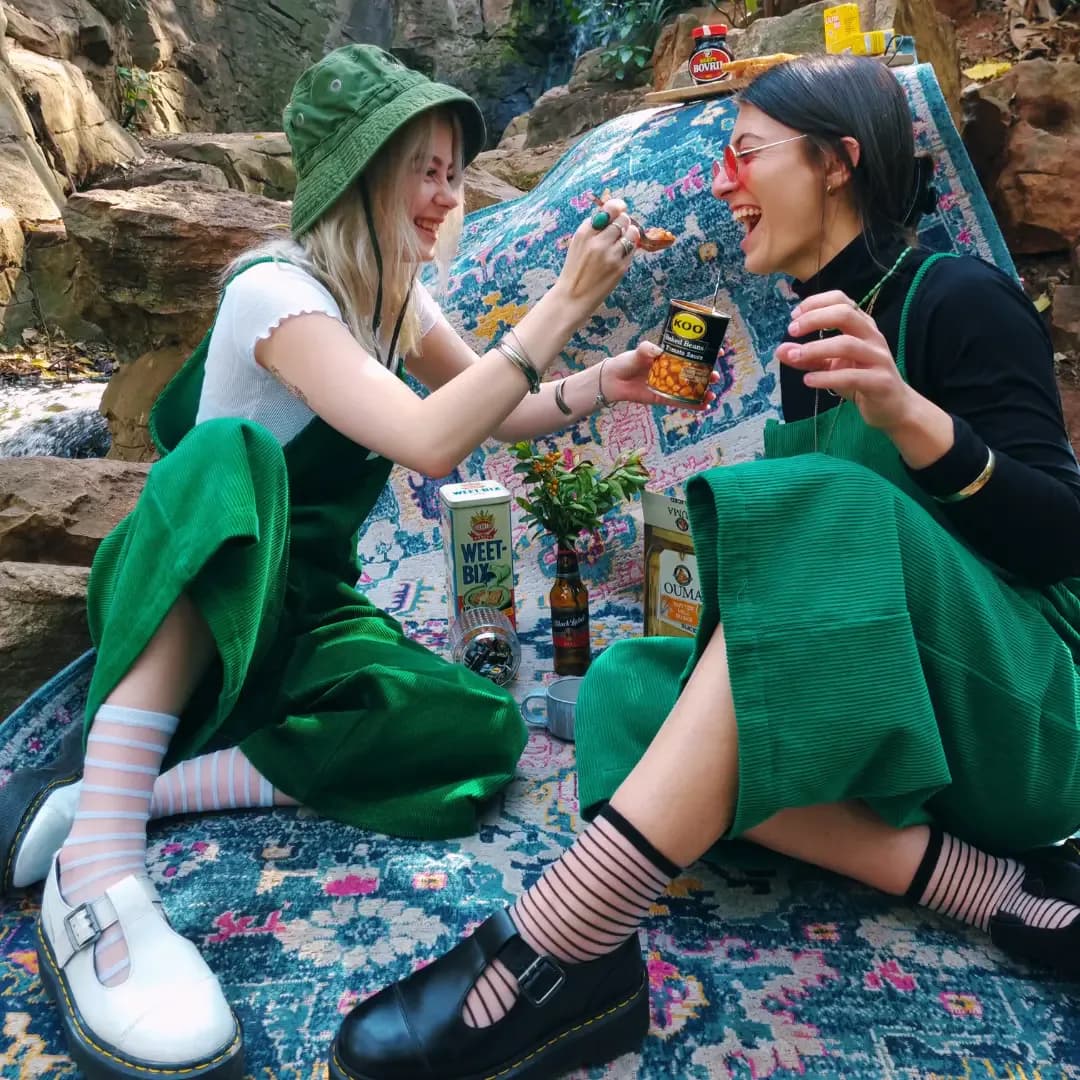 Two people in green PLEBS cotton corduroy dungarees sharing food at an outdoor picnic.